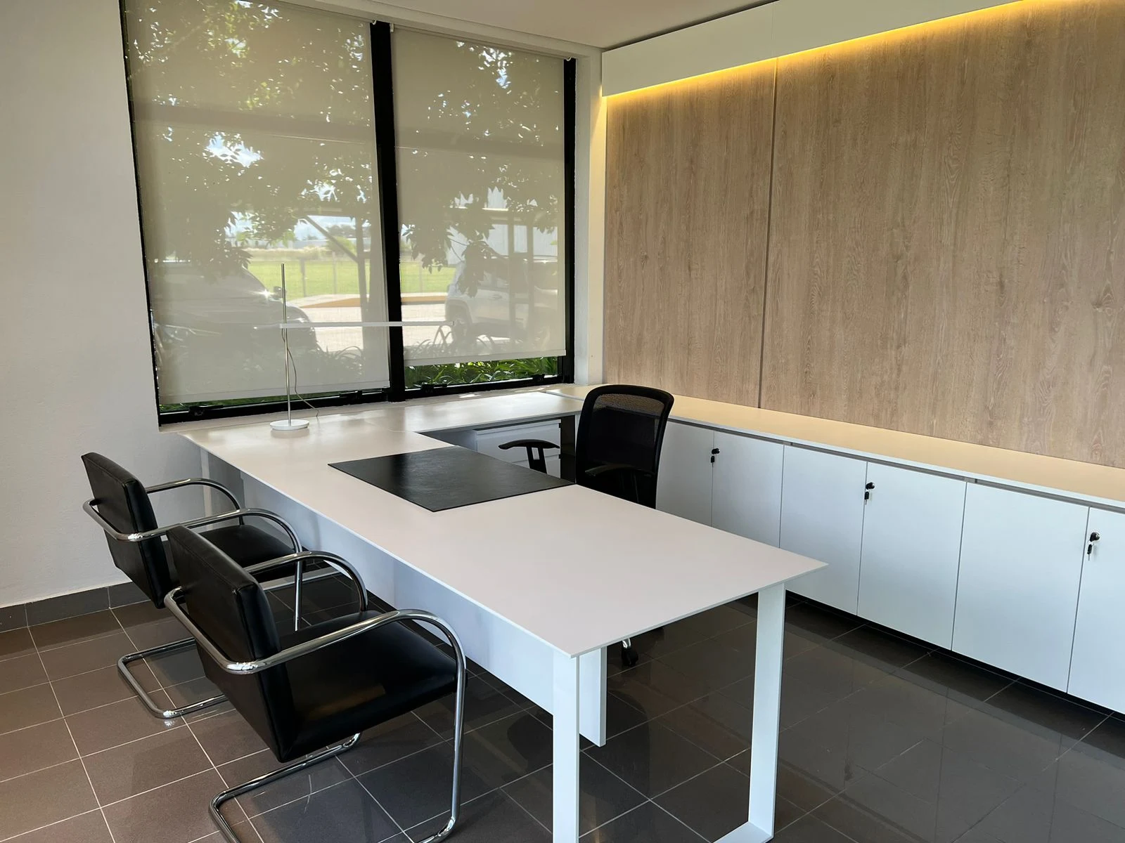 Despacho con escritorio claro, sillas de visita negras, guardado bajo y panel de tono madera iluminado junto a una ventana